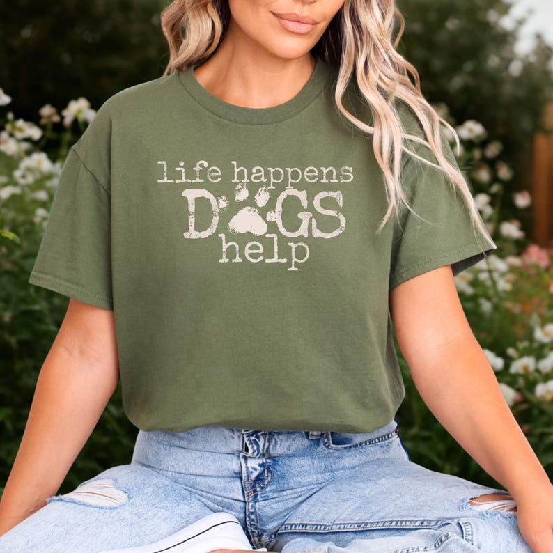 K&ouml;nnte beinhalten: Olivgr&uuml;nes T-Shirt mit dem Text "life happens DOGS help" in verblichener wei&szlig;er Schrift. Das Wort "DOGS" hat ein Pfotenabdruck-Design. Das Shirt ist kurz&auml;rmlig und hat einen Rundhalsausschnitt.