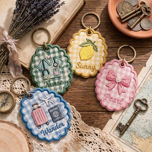 May include: Four embroidered keychains with scalloped edges and gold-tone rings. Designs include a nurse's uniform, a lemon with "Sunny" text, a pink bow with "Pearl" text, and a suitcase, airplane, and passport with "Wander" text. Each keychain has a gingham pattern.