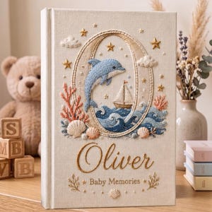 May include: Cream-colored baby memory book with an embroidered ocean scene featuring a blue dolphin, sailboat, and waves. The book is personalized with the name "Oliver" and the words "Baby Memories". Decorative elements include stars, clouds, and seashells.