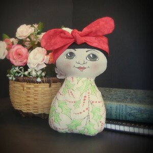 May include: A handmade doll with a red bow, green and white holly patterned dress, and a painted face. The doll is sitting in front of a basket of pink flowers.