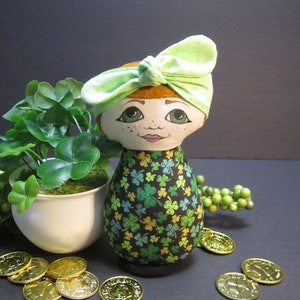 Op de afbeelding: Een groene en gouden stoffen pop met een groen hoofdband en klavertjevier print. De pop houdt een pot met klaverblaadjes vast en is omringd door gouden munten.