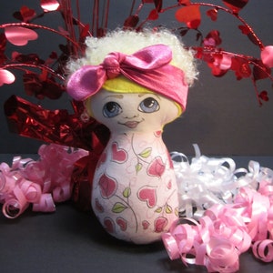 May include: A handmade doll with a heart-patterned dress, blonde curly hair, and a pink bow headband. The doll has a painted face with blue eyes and is surrounded by pink and white curly ribbons and red heart decorations.