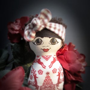 May include: A handmade doll with a white and red patterned dress, a red and white striped headband, and a painted face with brown eyes, red lips, and rosy cheeks.