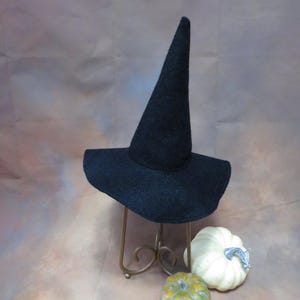May include: A black felt witch hat with a tall, pointed crown and a wide, slightly curved brim. The hat is displayed on a gold-toned metal stand, positioned next to one small, pale green pumpkin and one larger, white pumpkin with a textured surface.