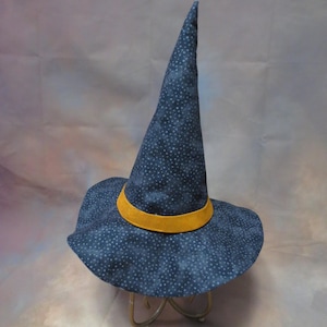May include: A pointed hat in a dark blue fabric featuring a small white star pattern. The hat includes a wide, flexible brim and a solid mustard yellow band encircling the base of the tall, conical crown, creating a distinct contrast.