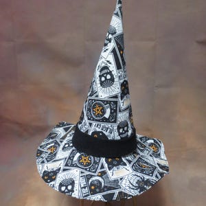 May include: A pointed witch hat featuring a black, white, and orange pattern of skulls, black cats, and tarot-style cards. The hat has a wide, slightly curved brim and a solid black fabric band encircling the base of the tall, conical crown.