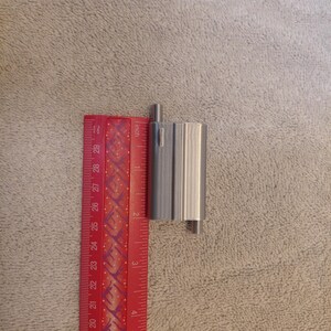 May include: A silver-colored, cylindrical metal object with a small, rectangular protrusion. The object is approximately 2 inches in height. A red ruler with inch markings is positioned next to the object, providing a size reference.