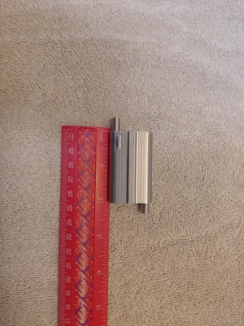 May include: A silver-colored, cylindrical metal object with a small, rectangular protrusion. The object is approximately 2 inches in height. A red ruler with inch markings is positioned next to the object, providing a size reference.