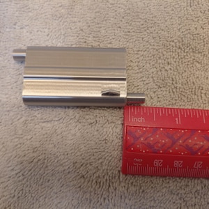 May include: A rectangular, brushed metal object with two cylindrical extensions, next to a red ruler. The ruler shows measurements in inches. The metal object appears to be approximately 2 inches long.