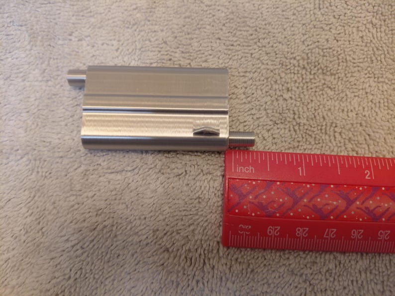 May include: A rectangular, brushed metal object with two cylindrical extensions, next to a red ruler. The ruler shows measurements in inches. The metal object appears to be approximately 2 inches long.