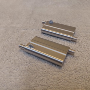 May include: Two brushed metal rectangular objects with cylindrical extensions on each end and a triangular detail on the top surface. The objects are lying on a light-colored, textured surface.