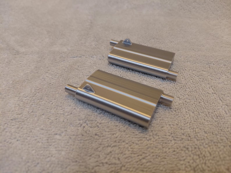 May include: Two brushed metal rectangular objects with cylindrical extensions on each end and a triangular detail on the top surface. The objects are lying on a light-colored, textured surface.