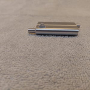 May include: A silver-colored metal door latch with a rectangular body and a small, cylindrical projection. The latch has a small, triangular loop on top. The object is resting on a textured, light gray surface.