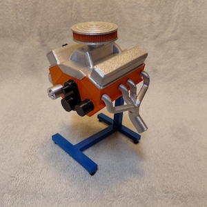 May include: A model engine, featuring a silver and orange color scheme, is mounted on a blue stand. The engine has a silver air filter and exhaust pipes. The stand is a simple, T-shaped design.