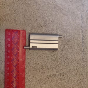 May include: A small, rectangular, silver-colored metal object with two thin, horizontal black lines and two cylindrical extensions. A red ruler with inch markings is next to the object.
