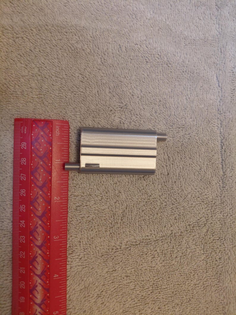 May include: A small, rectangular, silver-colored metal object with two thin, horizontal black lines and two cylindrical extensions. A red ruler with inch markings is next to the object.