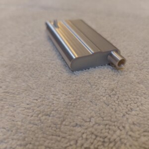 May include: A silver-colored metal object with a rectangular body and a cylindrical extension. The object has a smooth, reflective surface and appears to be a component or part of a larger mechanism. The object is resting on a textured, off-white surface.