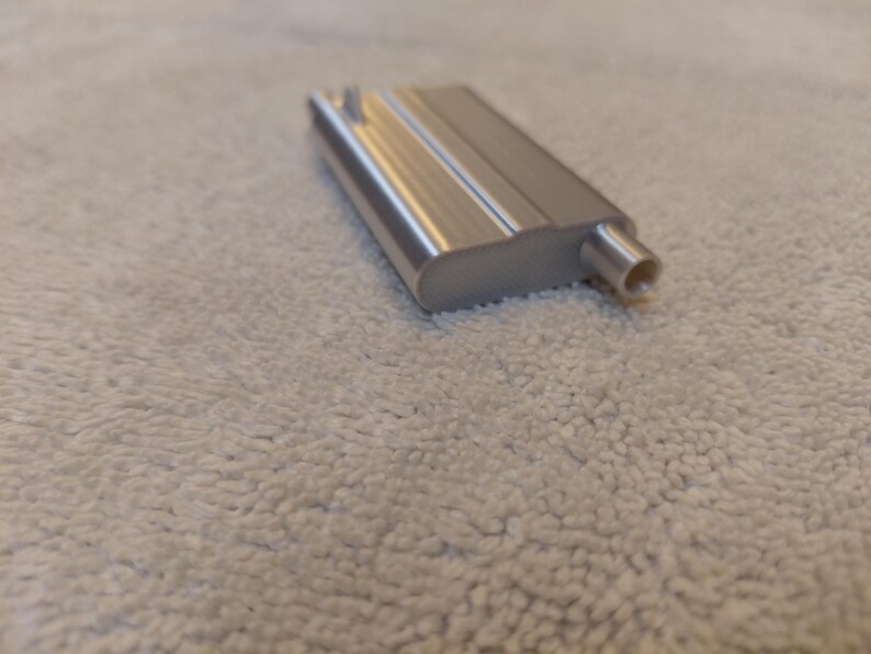 May include: A silver-colored metal object with a rectangular body and a cylindrical extension. The object has a smooth, reflective surface and appears to be a component or part of a larger mechanism. The object is resting on a textured, off-white surface.