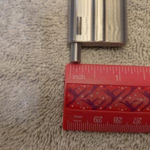 May include: A close-up of a silver metal object with a small cylindrical extension, next to a red ruler. The ruler shows the extension is approximately 0.25 inches long. The background is a textured, light gray surface.