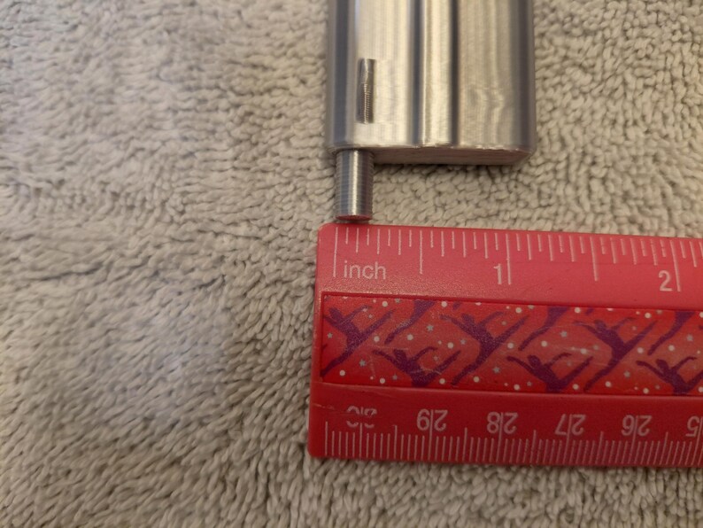 May include: A close-up of a silver metal object with a small cylindrical extension, next to a red ruler. The ruler shows the extension is approximately 0.25 inches long. The background is a textured, light gray surface.