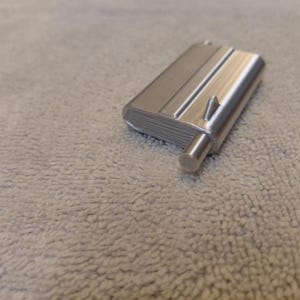 May include: A silver-colored, rectangular object with a cylindrical protrusion and a small, triangular detail. The object has a ridged surface and appears to be made of metal. The item is resting on a textured, light gray surface.