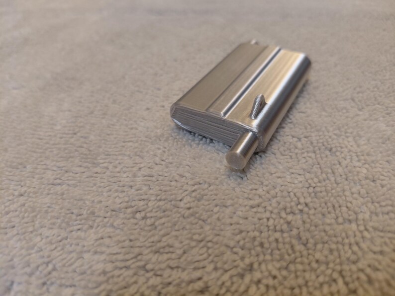 May include: A silver-colored, rectangular object with a cylindrical protrusion and a small, triangular detail. The object has a ridged surface and appears to be made of metal. The item is resting on a textured, light gray surface.