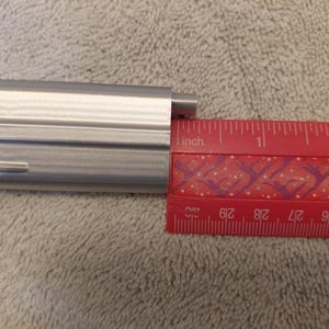 May include: A silver-colored metal object is positioned next to a red ruler. The ruler shows measurements in inches. The metal object appears to be approximately 1.5 inches long.