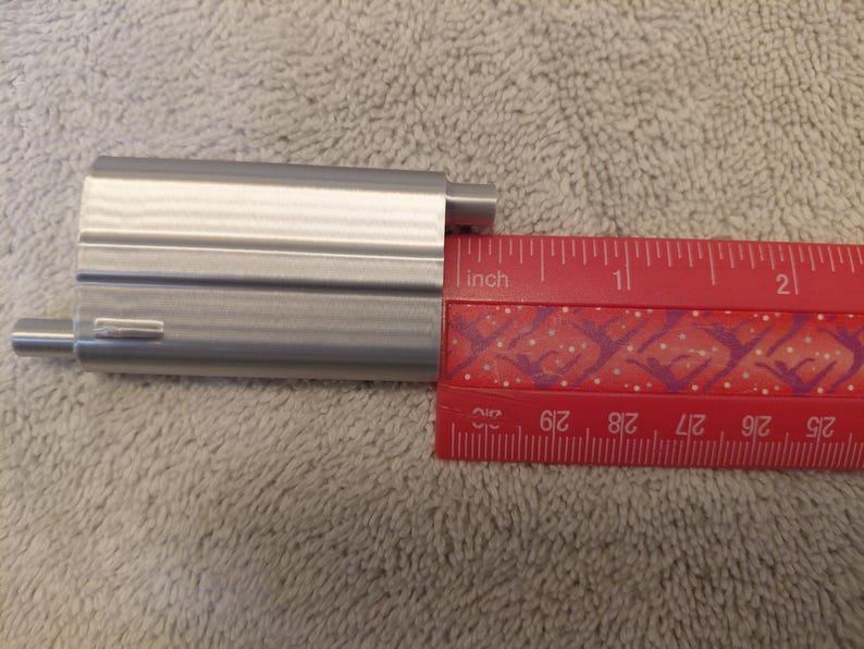 May include: A silver-colored metal object is positioned next to a red ruler. The ruler shows measurements in inches. The metal object appears to be approximately 1.5 inches long.