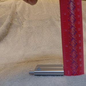 May include: A red ruler with white markings is positioned next to a small, silver metal object. The ruler shows measurements in inches. The object appears to be a tool or part of a larger item, resting on a textured, off-white surface.