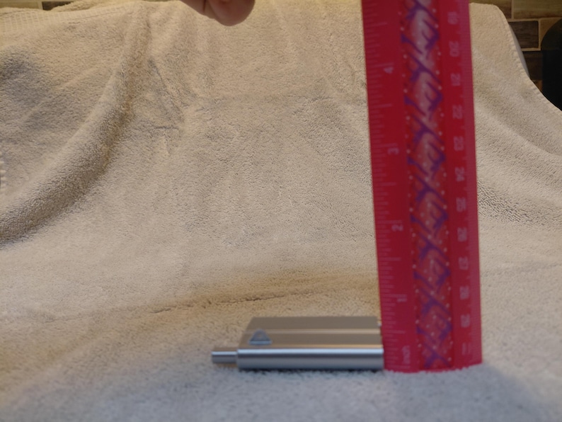May include: A red ruler with white markings is positioned next to a small, silver metal object. The ruler shows measurements in inches. The object appears to be a tool or part of a larger item, resting on a textured, off-white surface.