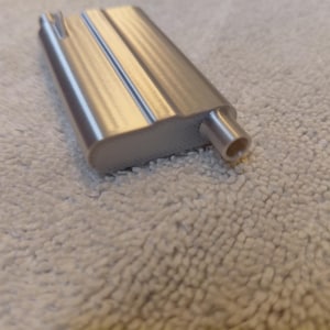 May include: A silver-colored, rectangular metal object with a rounded edge and a cylindrical protrusion. The object has a textured surface and appears to be a component or part of a larger device. The background is a light-colored, textured fabric.