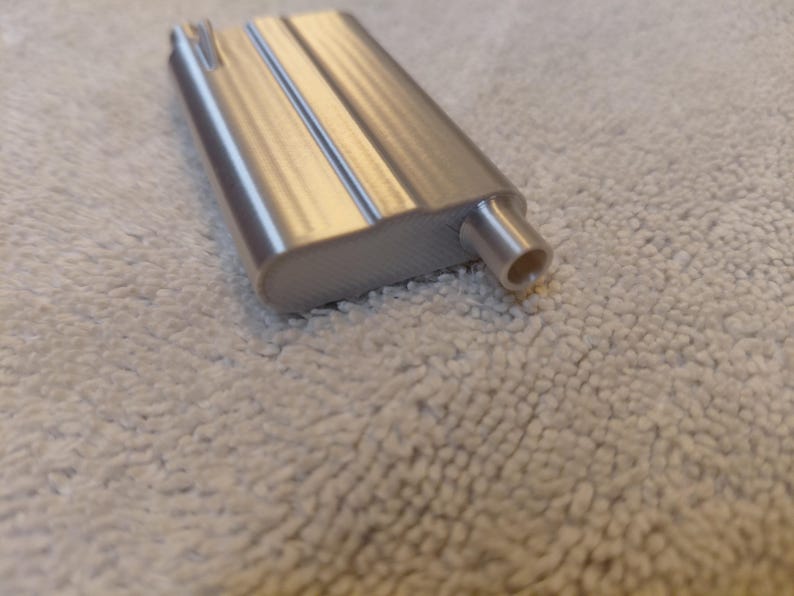 May include: A silver-colored, rectangular metal object with a rounded edge and a cylindrical protrusion. The object has a textured surface and appears to be a component or part of a larger device. The background is a light-colored, textured fabric.