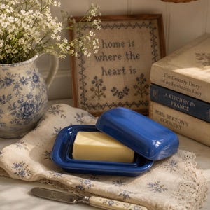 May include: A glossy cobalt blue ceramic butter dish with a matching lid, containing a rectangular block of yellow butter. The dish rests on a cream-colored fabric napkin with a blue floral pattern, accompanied by a silver butter knife with a floral handle.