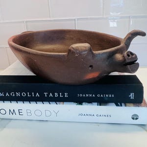 Puede incluir: Un cuenco marrón de cerámica con forma de cerdo, de forma ovalada y ancha. El cuenco tiene hocico, orejas y cola enroscada. Descansa sobre dos libros, uno titulado "Magnolia Table" y otro "Somebody" de Joanna Gaines.