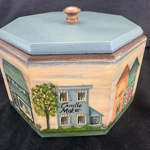 Puede incluir: Caja hexagonal de madera pintada a mano con tapa. La caja presenta escenas pintadas de tiendas, incluyendo un "Candle Maker" y un "Coffee Shop". La tapa es de un azul verdoso apagado y el pomo es marrón.