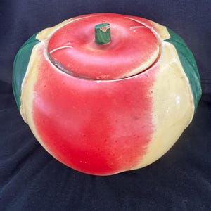 Può includere: Un barattolo per biscotti in ceramica a forma di mela con coperchio. Il barattolo è principalmente rosso con accenti gialli e verdi, imitando l'aspetto di una vera mela. Il coperchio ha un gambo verde.