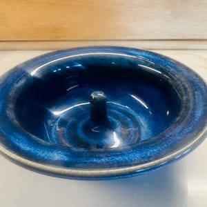 Puede incluir: Un plato circular de cerámica para anillos con un esmalte azul profundo y variaciones tonales en azul claro y negro. El cuenco tiene un acabado liso y brillante, con un poste central vertical para colocar anillos y un borde ancho y redondeado.