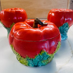 Pode incluir: Um conjunto de três itens de cozinha em cerâmica com formato de tomate vermelho e base de folhas verdes. Dois itens são saleiros altos com pequenos orifícios no topo e um é um recipiente redondo com tampa e um puxador de caule castanho.