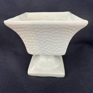 Pode incluir: Uma tigela de pedestal de cerâmica branca com uma superfície texturizada e com covinhas. A tigela tem uma base quadrada e um topo hexagonal alargado. O design é simples e elegante, adequado para fins decorativos.