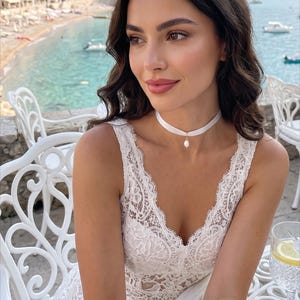 May include: A woman in a white lace dress and a white choker necklace with a pearl pendant. She is seated at a white table with a glass of water and lemon. The background features a coastal scene with buildings and boats.