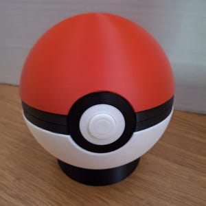 May include: A spherical object featuring a red top half and a white bottom half, separated by a thick black horizontal band. A circular black and white button is centered on the front of the sphere, which rests on a small black circular base.