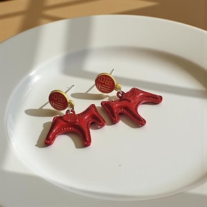 May include: A pair of red horse-shaped earrings with gold-coloured studs. The earrings are on a white plate. The earrings have a circular top with Chinese characters. The horse-shaped part of the earrings are a bright red colour.