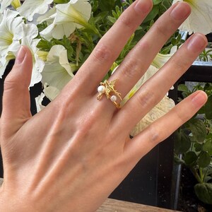 May include: A gold-colored ring featuring a bird design with two small, round, white pearls. The ring is on a hand with a wooden table and white flowers in the background. The ring is a piece of jewelry.
