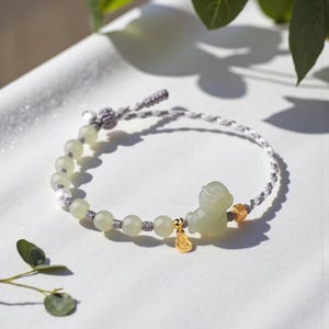 Peut inclure: Un bracelet de perles vert clair avec une petite figurine de jade et des accents dorés. Le bracelet est composé d'un cordon tressé blanc et gris et orné d'un petit charme doré. Les perles rondes sont espacées uniformément.