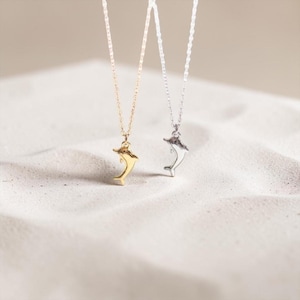 May include: Two dolphin pendant necklaces. One is gold-toned, the other silver-toned, both suspended on delicate chains. The pendants are detailed with a sleek, curved design. The necklaces rest on a textured, sandy surface, creating a beachy aesthetic.