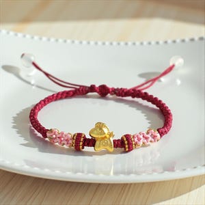 Può includere: Un braccialetto rosso intrecciato con un ciondolo dorato al centro. Il braccialetto presenta dettagli rosa e oro. Il cordino regolabile ha perline trasparenti alle estremità. Il braccialetto è esposto su un piatto bianco.