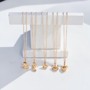 May include: Gold-colored necklaces with ocean-themed pendants, including an octopus, seahorse, fish, crab, and turtle. The necklaces hang from a white display stand against a white background. Each pendant is detailed and hangs from a delicate chain.