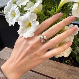 May include: A gold ring with a floral design, featuring a pearl, a red gemstone, and small turquoise stones. The ring is on a hand, with white petunia flowers in the background.