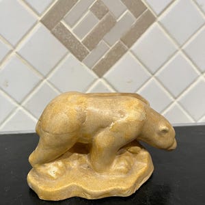 May include: A tan-colored bear figurine in a walking pose, sculpted with a smooth, matte finish. The bear is depicted in a low-slung stance with its head lowered, standing on an integrated, irregularly shaped base that mimics a rocky surface.