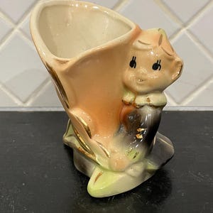 May include: A ceramic planter shaped like a cornucopia with a small figure attached to the side. The piece features a peach-colored horn, a figure with a light-colored face and dark clothing, and gold-painted accents on the edges and details.